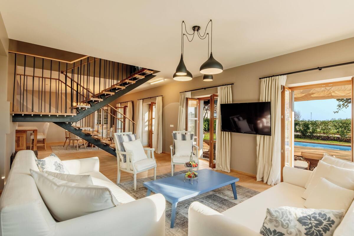 A spacious living area with a staircase, white furniture, a blue coffee table, and a TV.