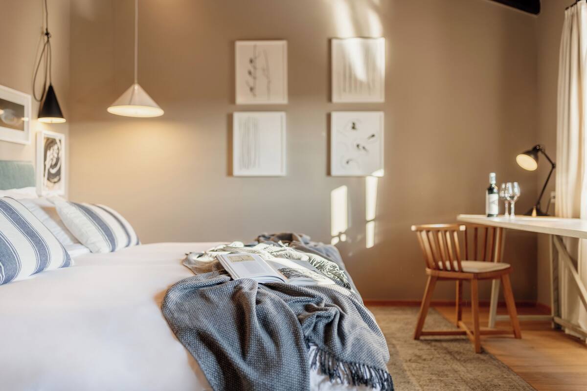 A neatly made bed with a blue and white striped pillow, a blue blanket, and a book on it. There is a wooden chair and a small table with a lamp and a bottle on it.