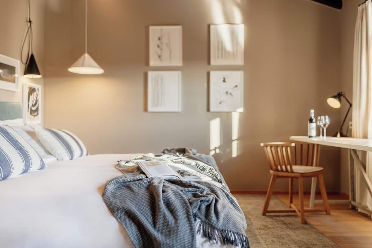 Una cama bien tendida con un cojín azul y blanco a rayas, una manta azul y un libro encima. Hay una silla de madera y una mesita con una lámpara y una botella.