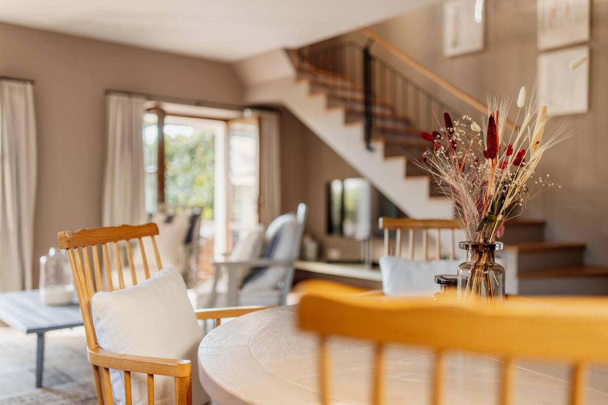 A dining area with a wooden table and chairs, a vase with dried flowers, and a staircase in the background.