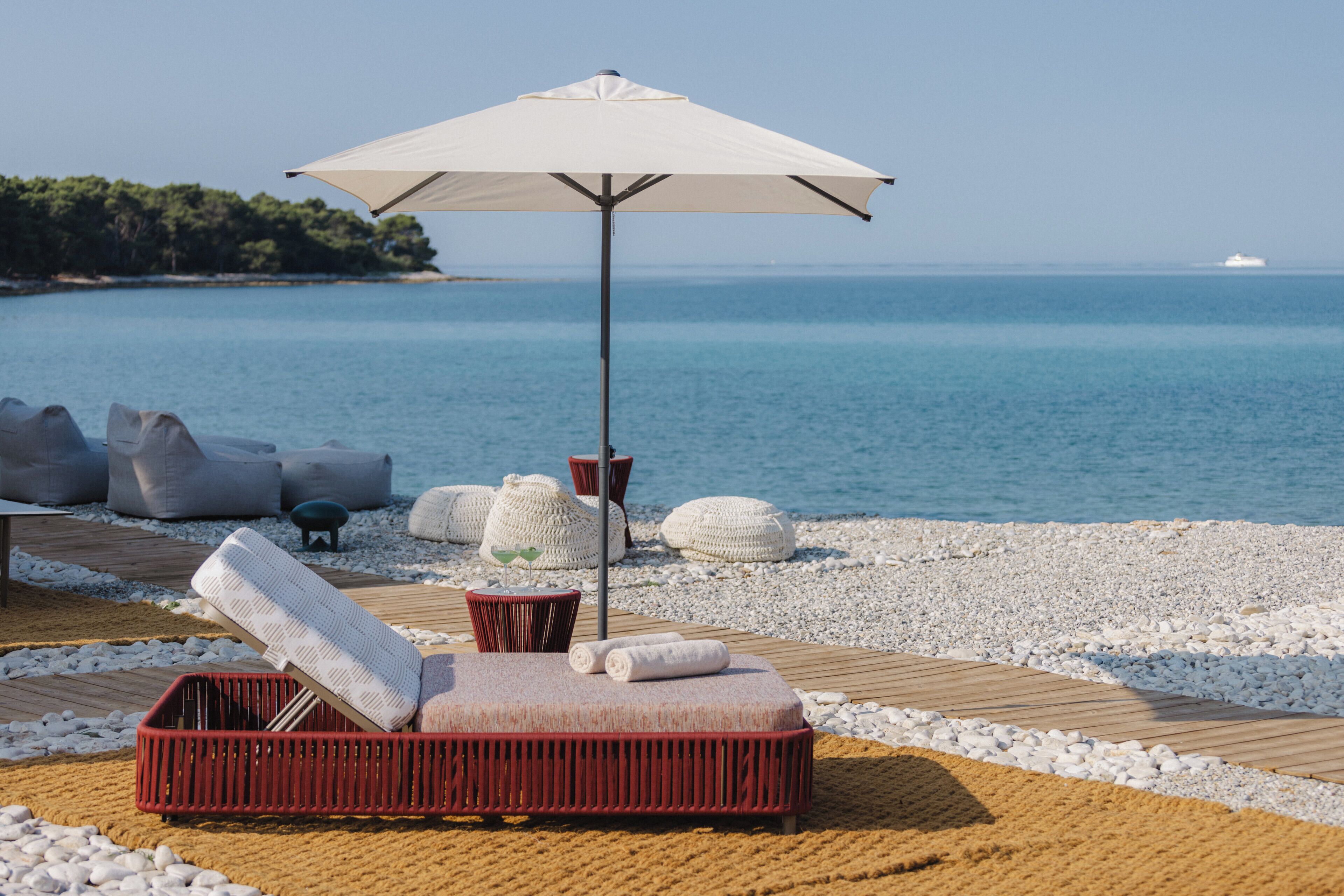 Un salón junto a la playa con una silla reclinable roja, una sombrilla blanca y una terraza de madera.