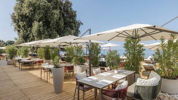 An outdoor dining area with wooden tables, chairs, and umbrellas, surrounded by potted plants and trees.