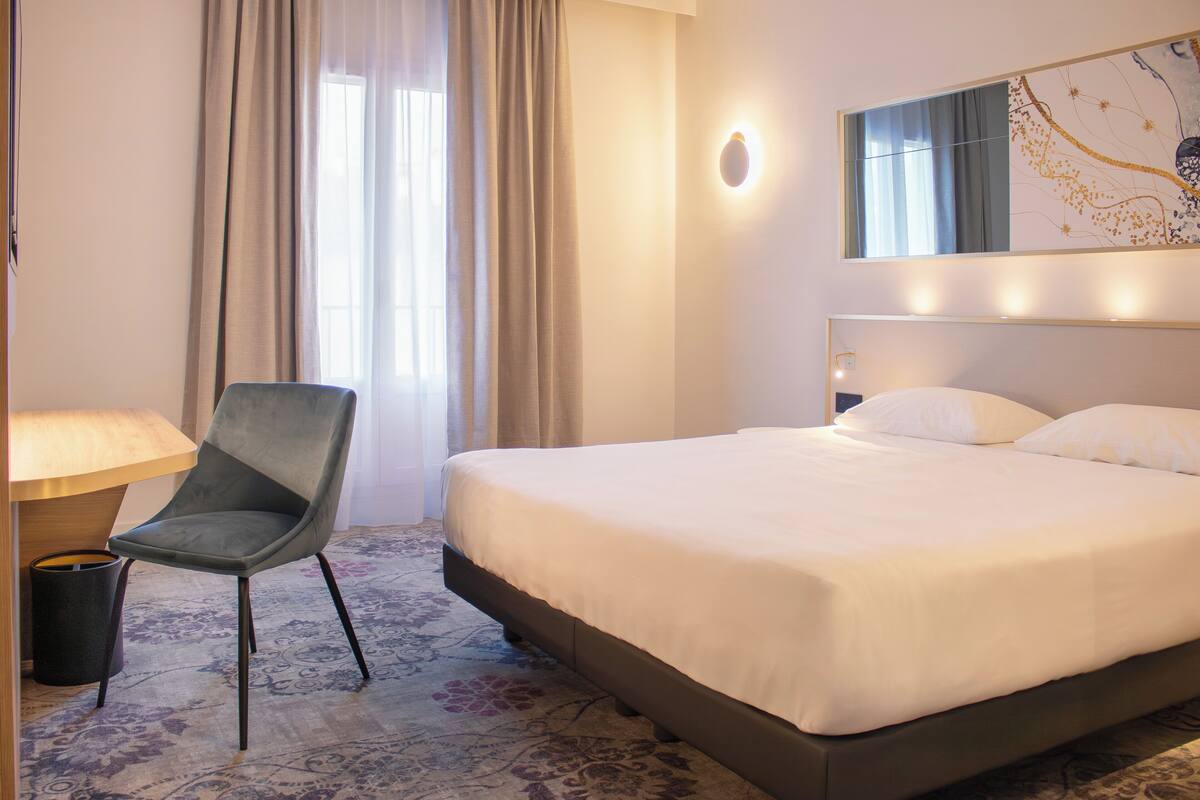 Ein Hotelzimmer mit einem Bett, einem Sessel, einer Lampe und einem großen Fenster mit Vorhängen.