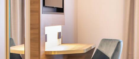 A hotel room with a wooden desk, a modern chair, a flat-screen TV mounted on the wall, and a patterned carpet.