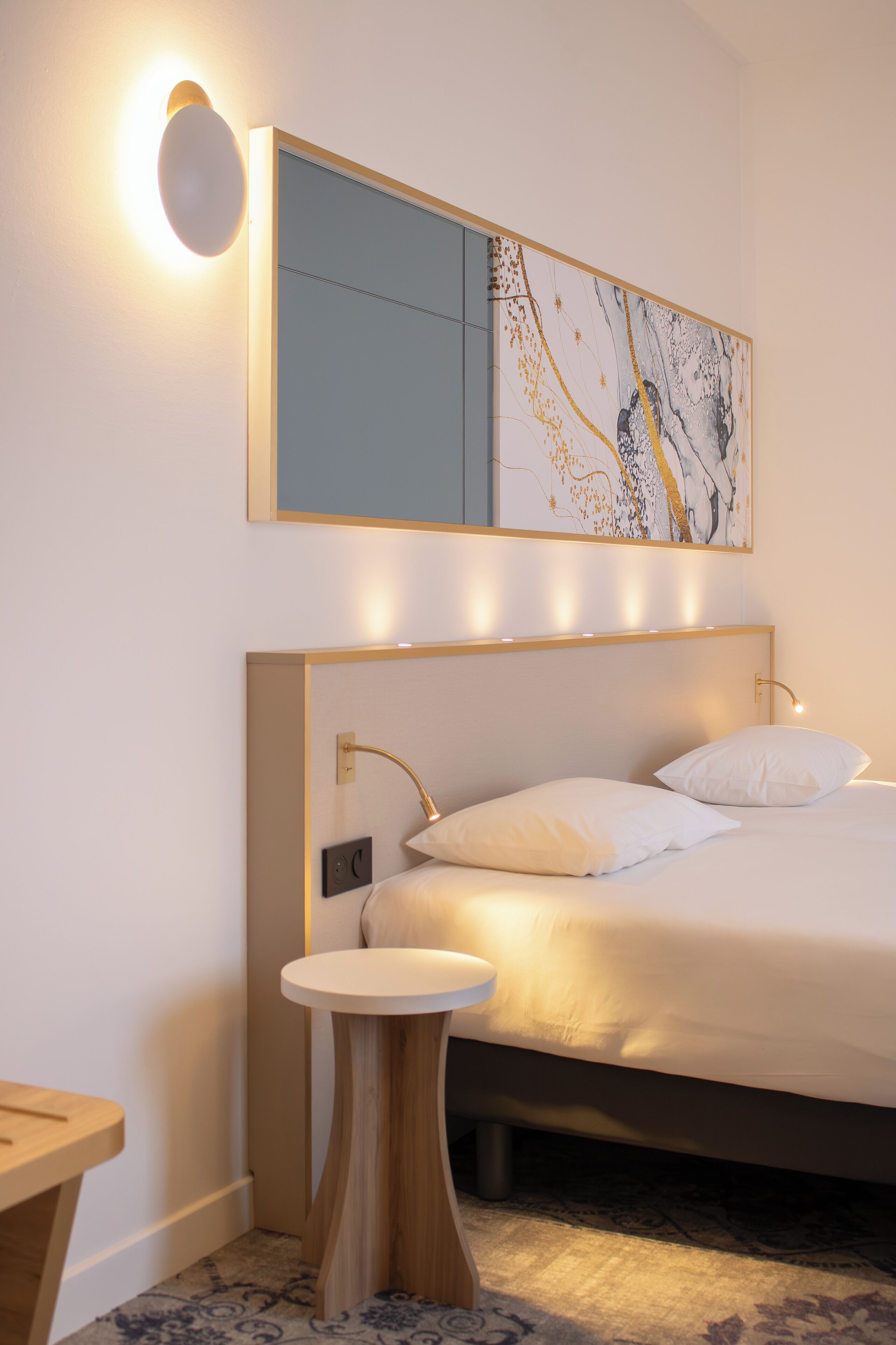 A modern hotel room with a large bed, bedside table, and a wall-mounted light fixture.
