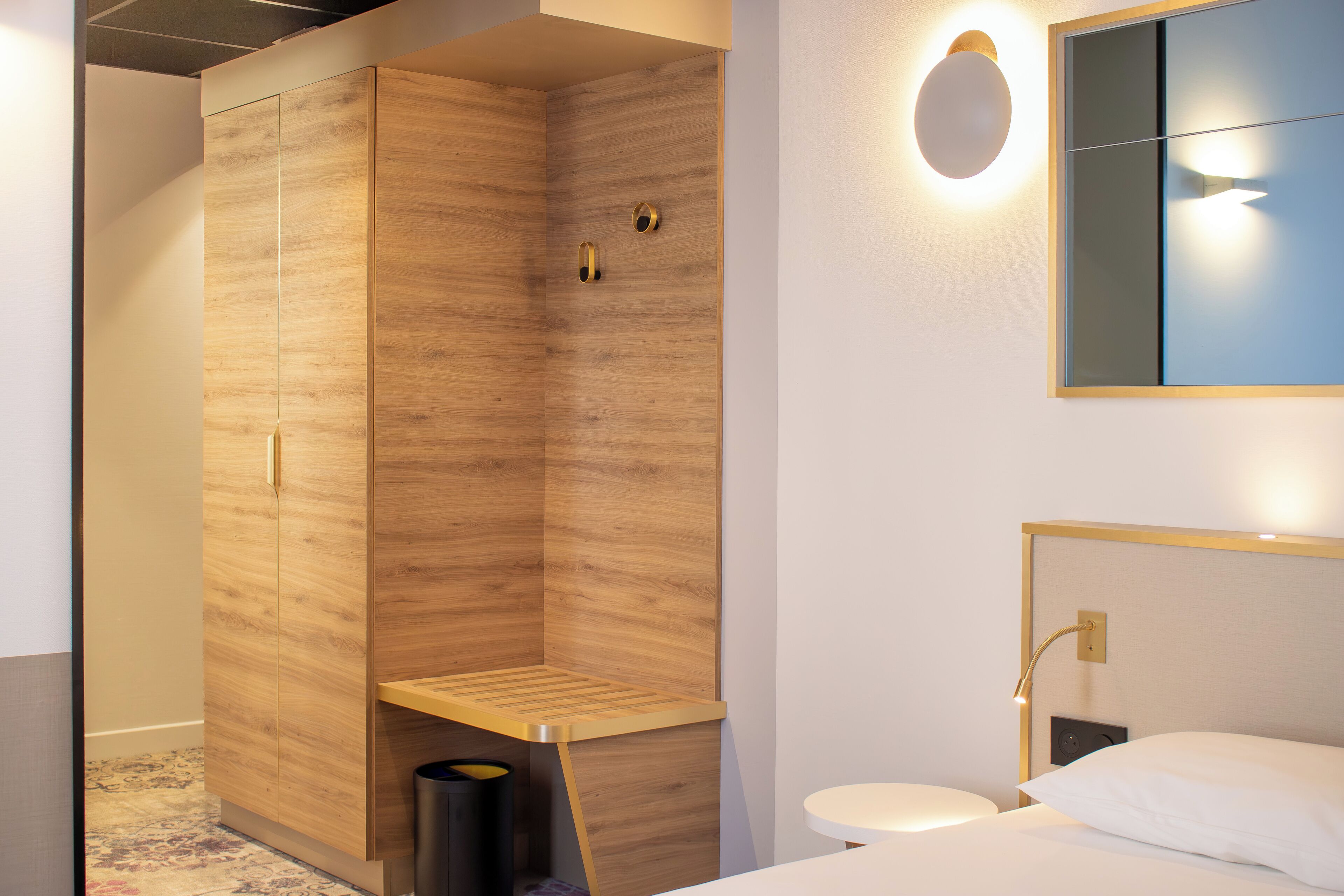 Una habitación de hotel moderna con un armario de madera, un pequeño banco, un cesto de basura, un espejo redondo, una mesita de noche y una cama con ropa blanca.