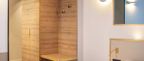 Una habitación de hotel moderna con un armario de madera, un pequeño banco, un cesto de basura, un espejo redondo, una mesita de noche y una cama con ropa blanca.