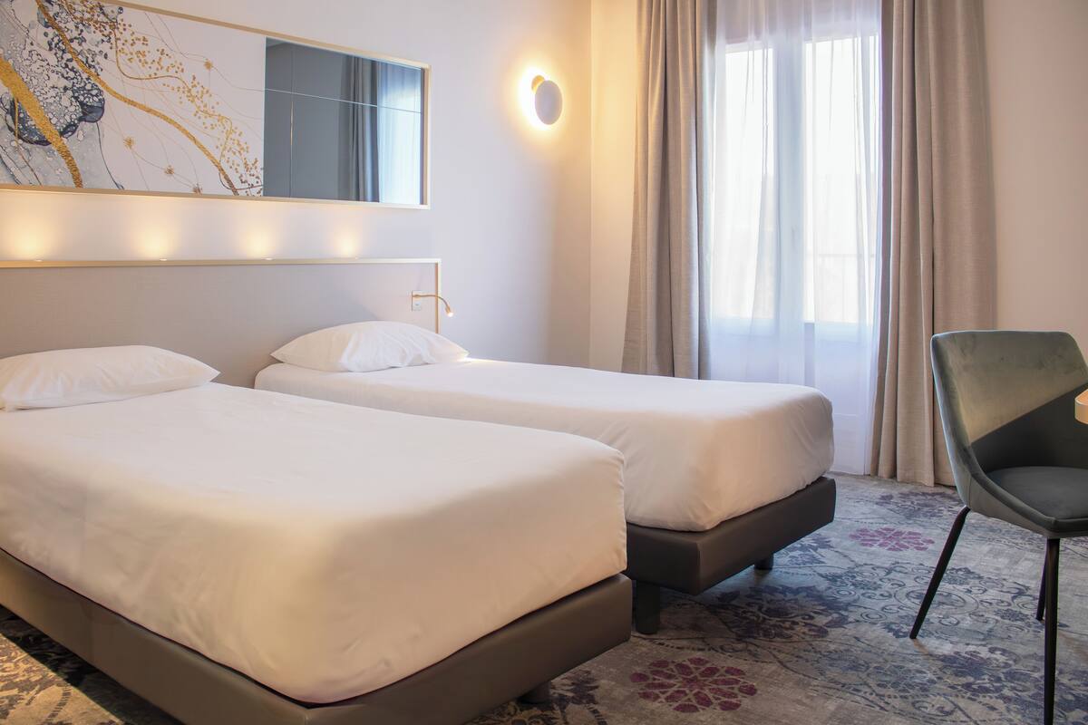 Ein Hotelzimmer mit zwei Betten, einem Sessel, einem großen Spiegel und einem Fenster mit Vorhängen.