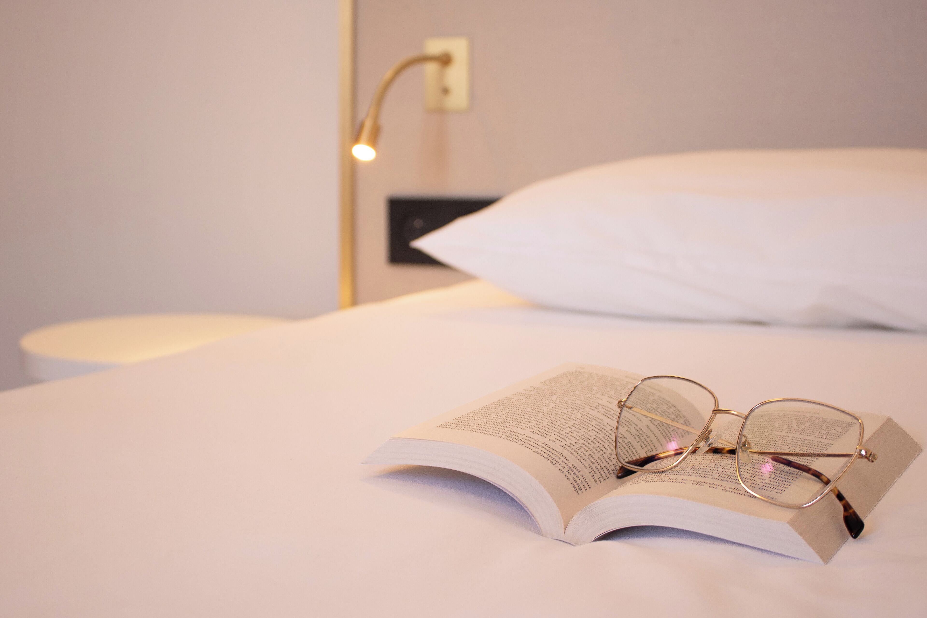 Una cama bien hecha con un almohadón blanco y un libro abierto con gafas encima.