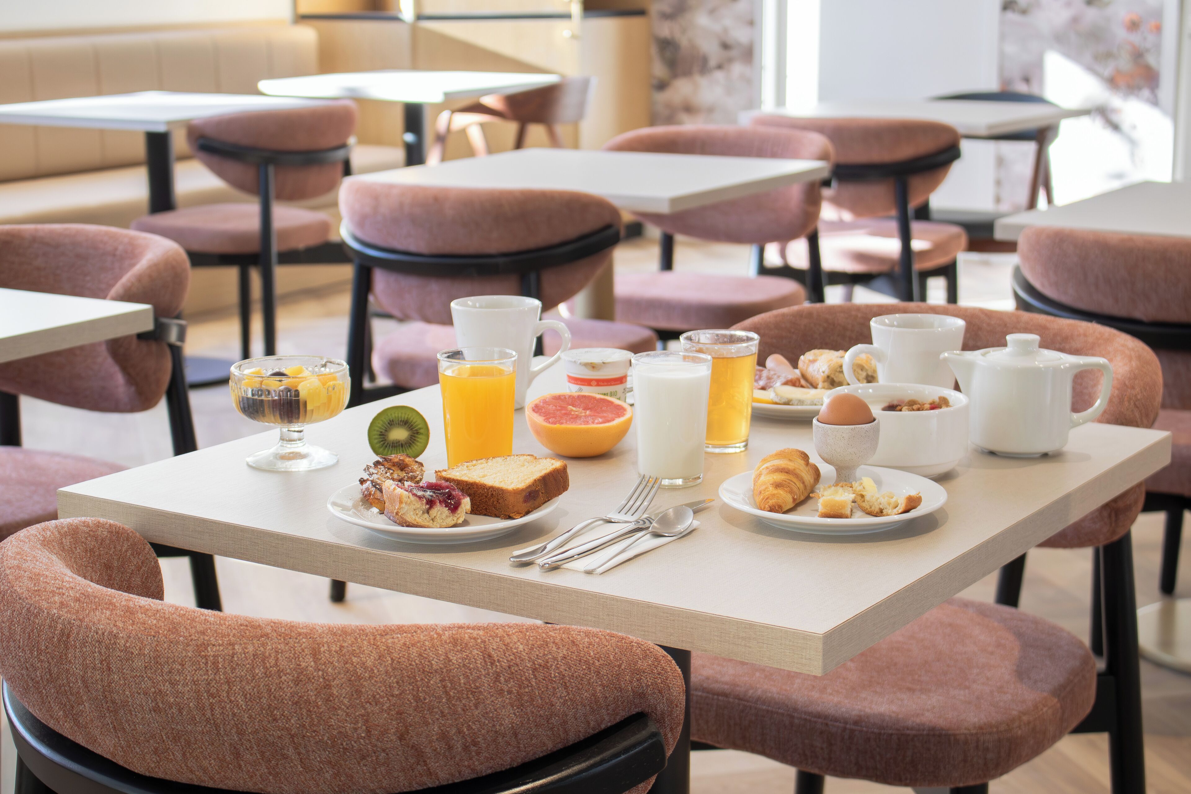 A breakfast setting with a table set for two, featuring plates of toast, fruit, and pastries, along with a jug of orange juice and a milk jug.