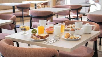 Une table mise pour le petit-déjeuner, avec une nappe et de la vaisselle pour deux personnes, comprenant des assiettes de toasts, des fruits et des viennoiseries, ainsi qu'une carafe de jus d'orange et un pot à lait.
