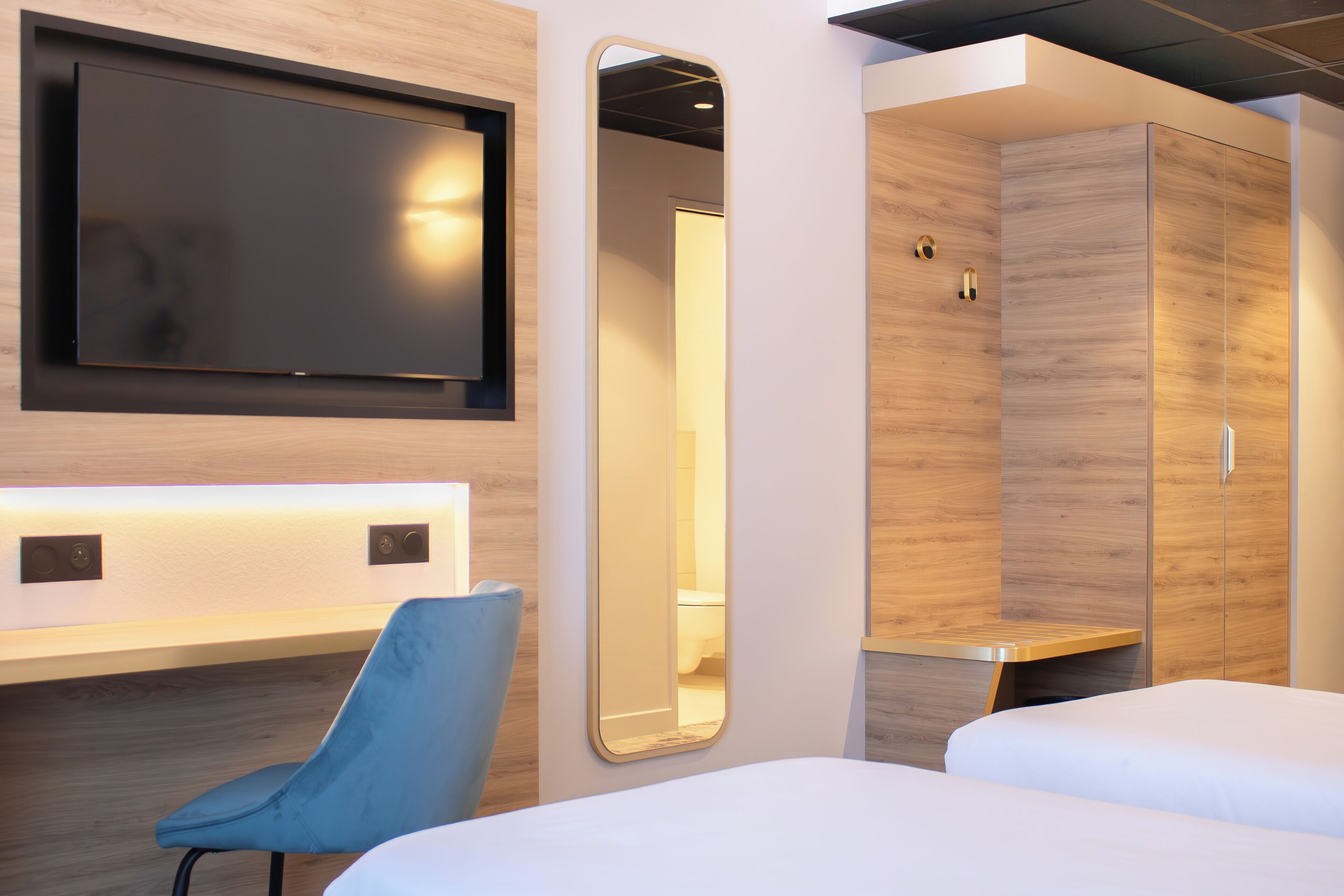 A modern hotel room with a flat-screen TV mounted on the wall, a large mirror, a wardrobe, and a blue chair.