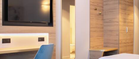 A modern hotel room with a flat-screen TV mounted on the wall, a large mirror, a wardrobe, and a blue chair.