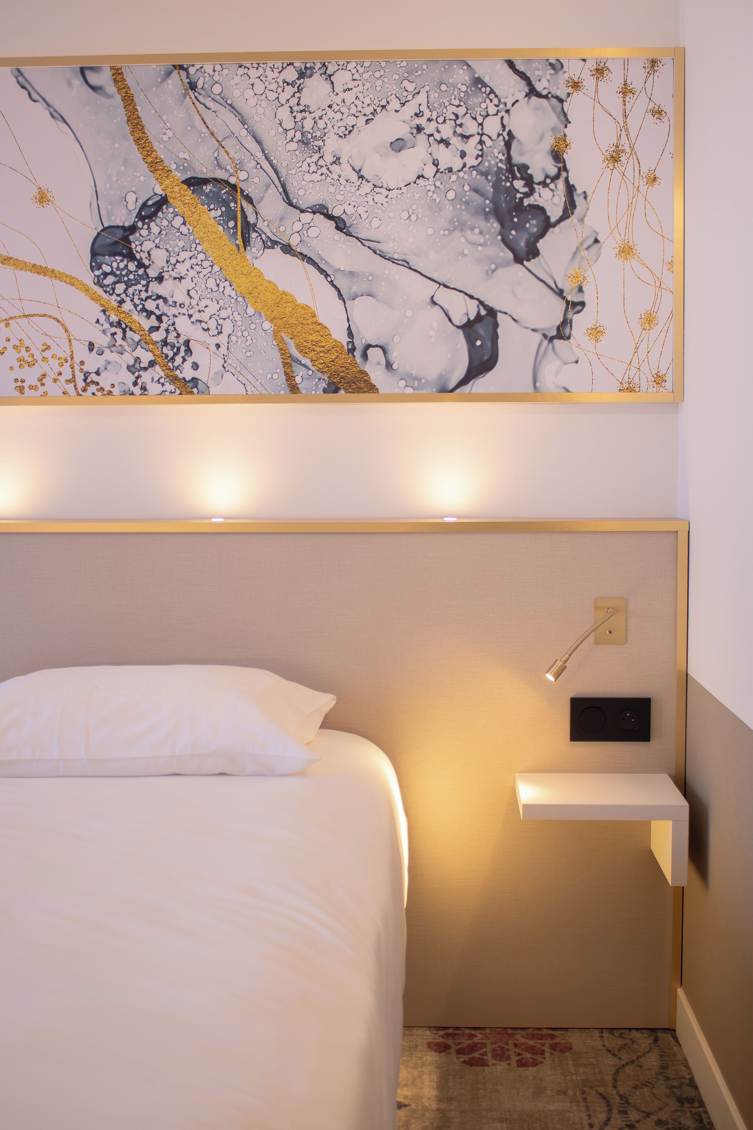 A hotel room with a bed, a bedside table, a wall-mounted lamp, and a decorative painting above the bed.