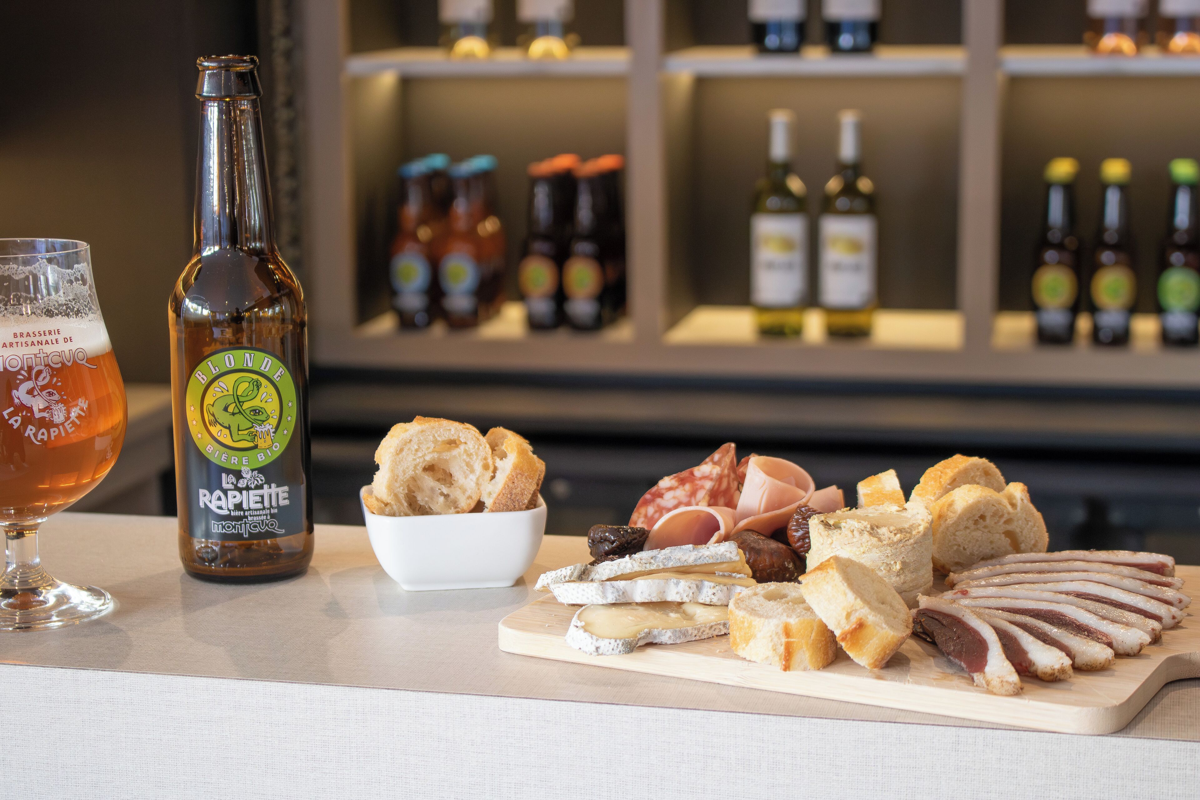 A bottle of blonde beer, a glass of beer, and a variety of cheeses and meats on a wooden board.