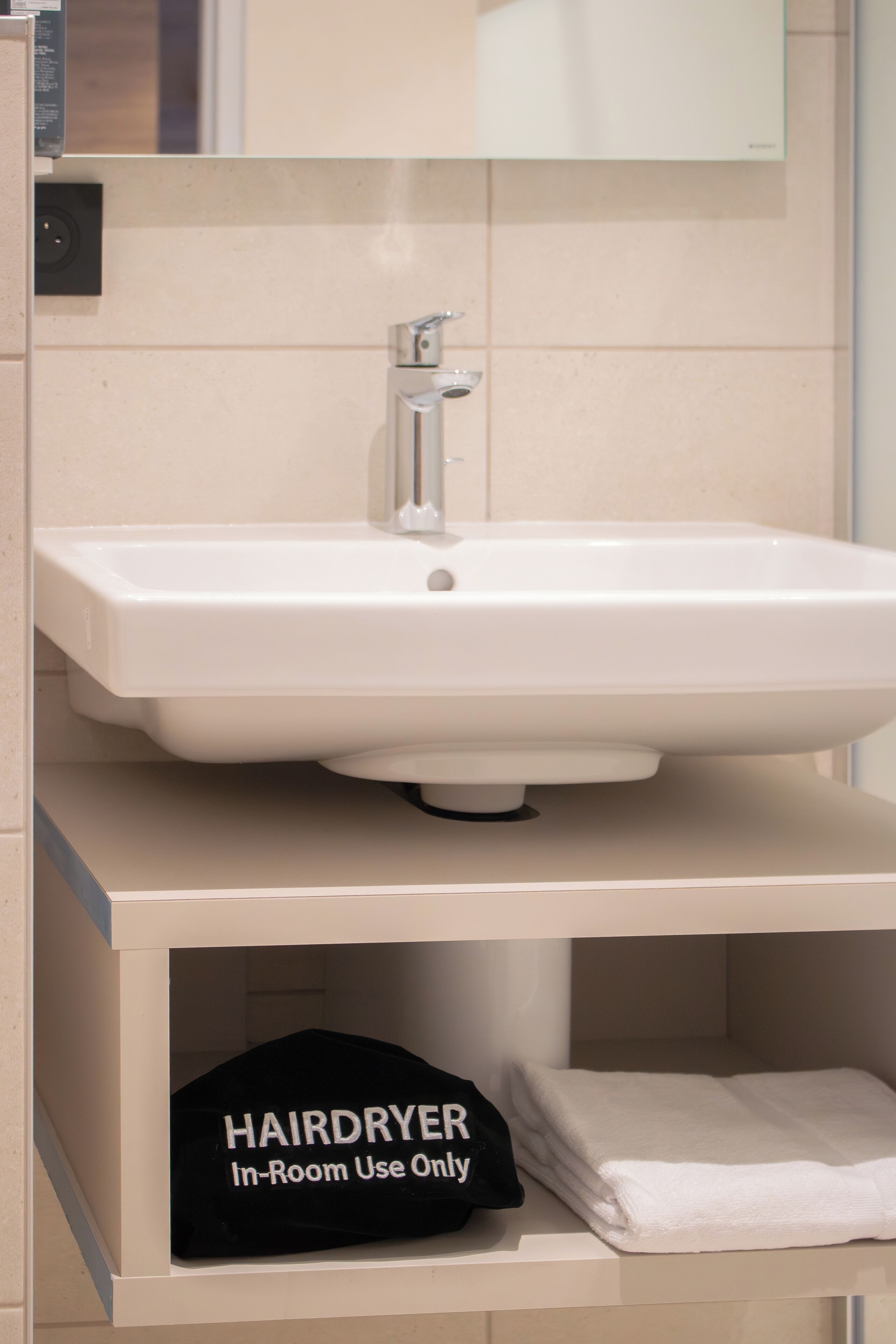 Un baño con un lavabo blanco, un espejo encima y un estante abajo con una gorra negra y una toalla blanca doblada.