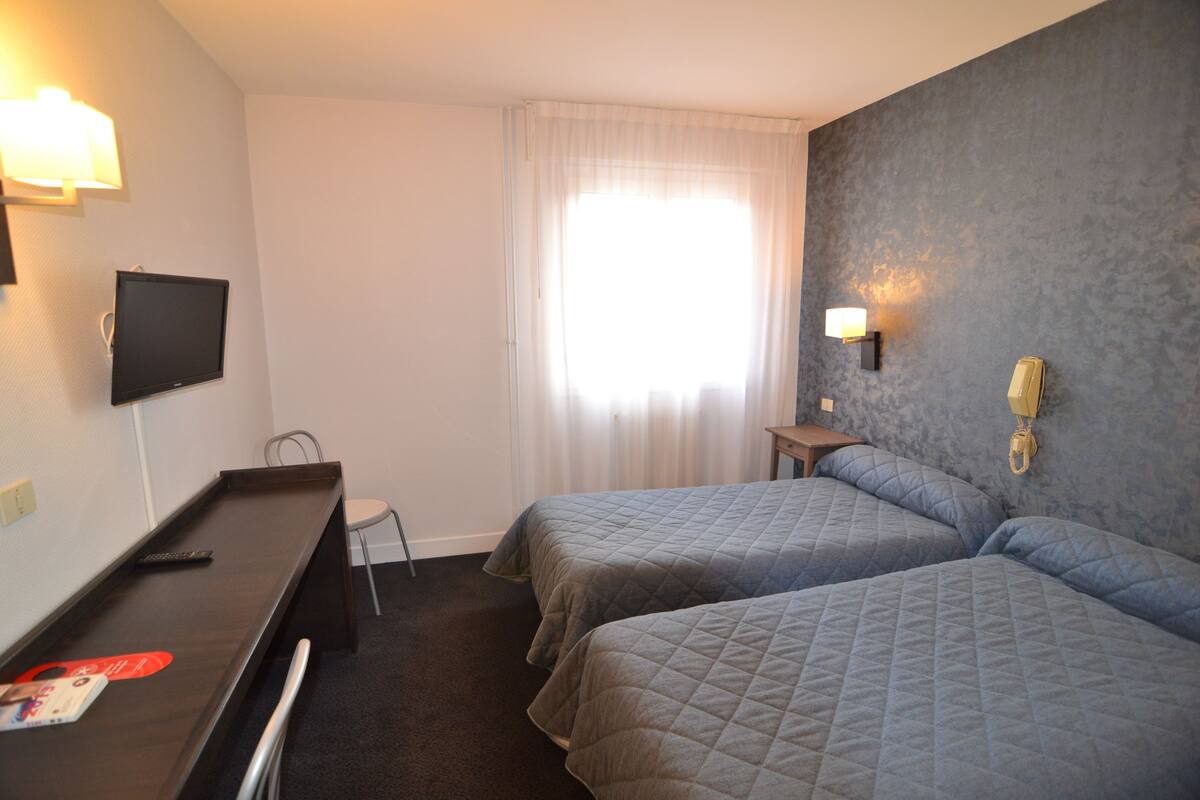 Ein Hotelzimmer mit zwei Betten, einem Schreibtisch, einem Fernseher und einem Fenster mit Vorhängen.