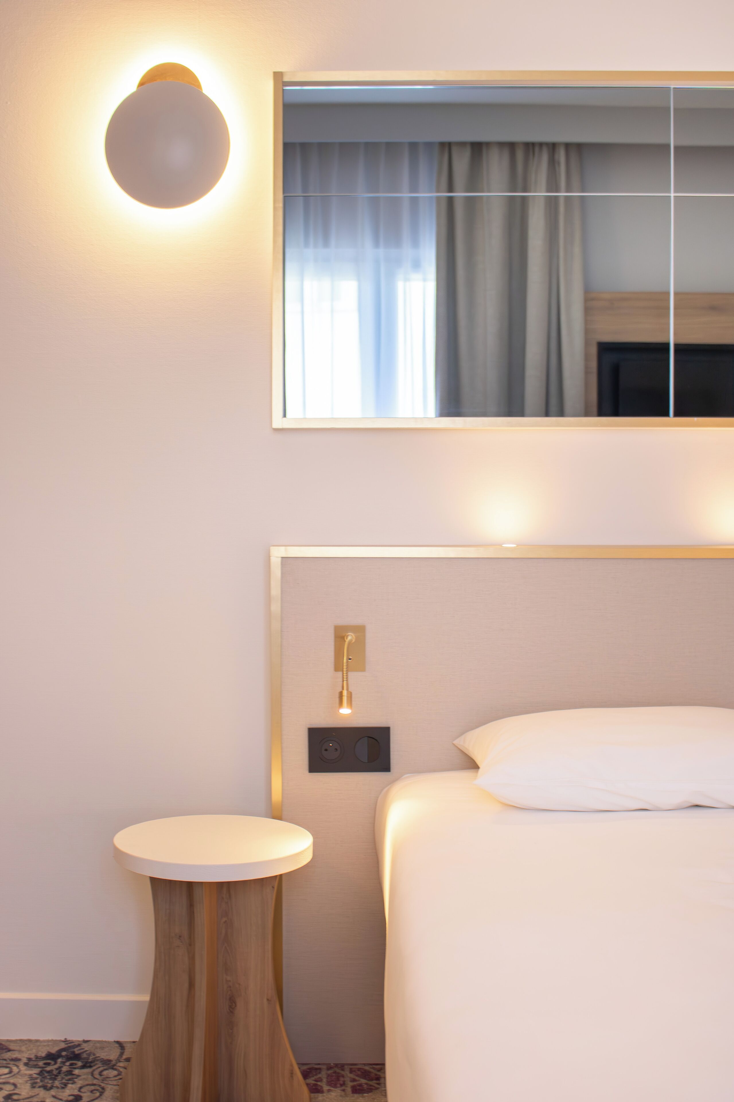 Une chambre d’hôtel avec un lit, une petite table ronde, une lampe fixée au mur et un miroir qui reflète une fenêtre avec des rideaux.
