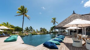 Una terraza de madera con sillones de descanso y sombrillas, una piscina y palmeras.