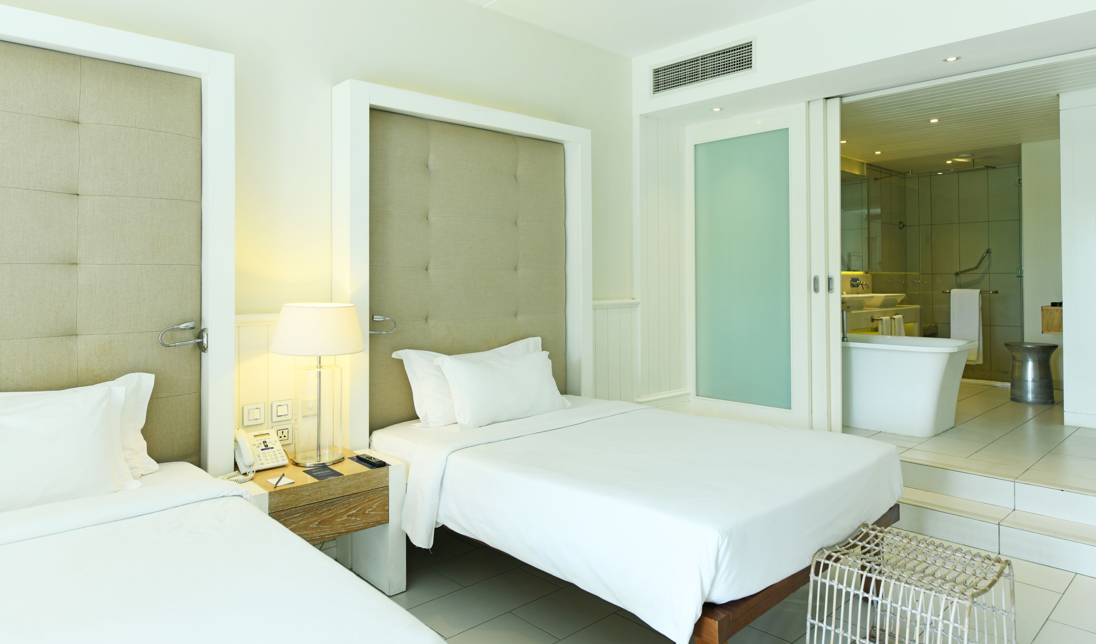 A hotel room with two beds, a bedside table with a lamp, a phone, and a small basket. There is a bathroom visible through an open door with a shower, sink, and toilet.