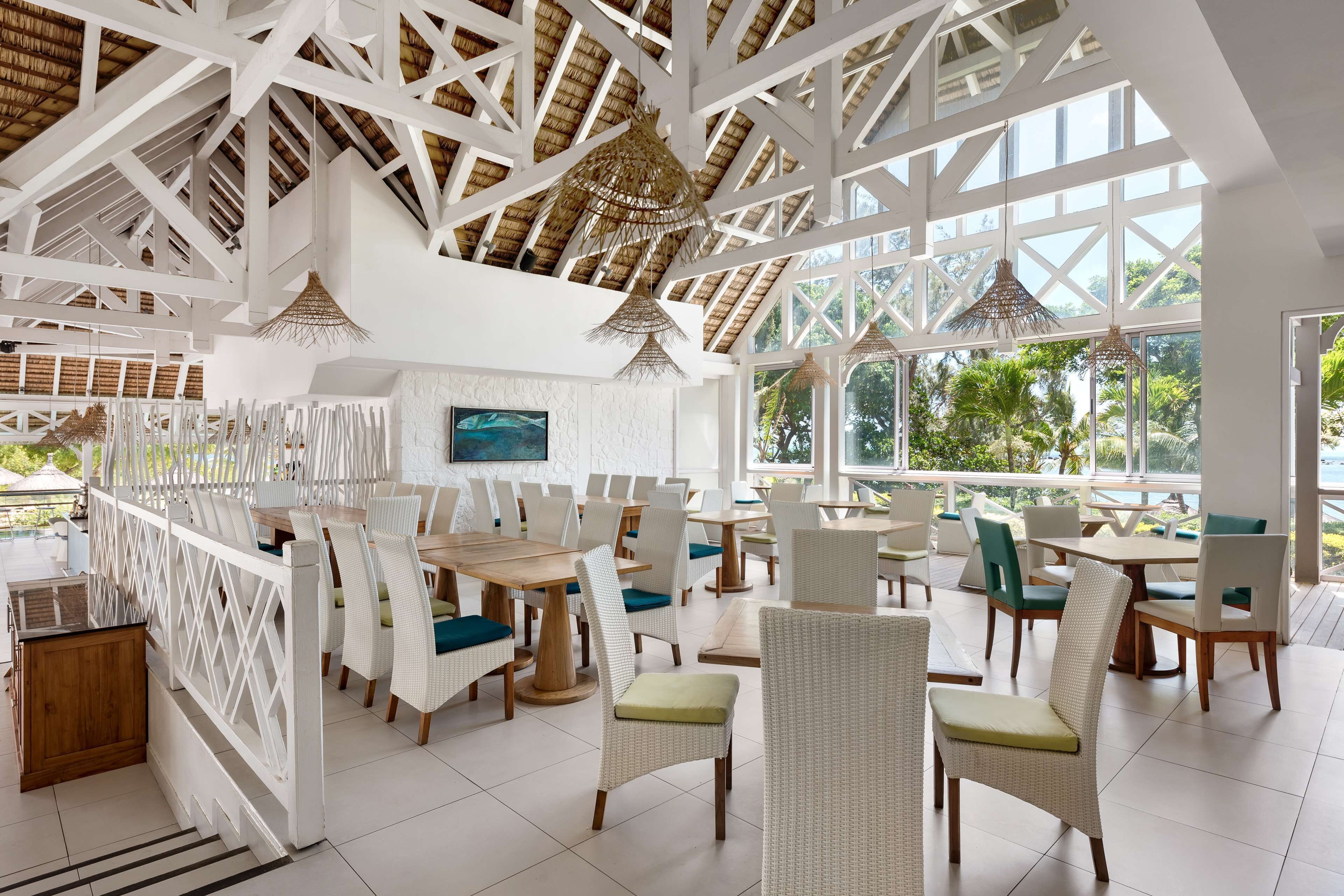 Un vaste espace intérieur aménagé en salle à manger, avec des meubles blancs, de grandes fenêtres et un plafond haut orné de poutres en bois.