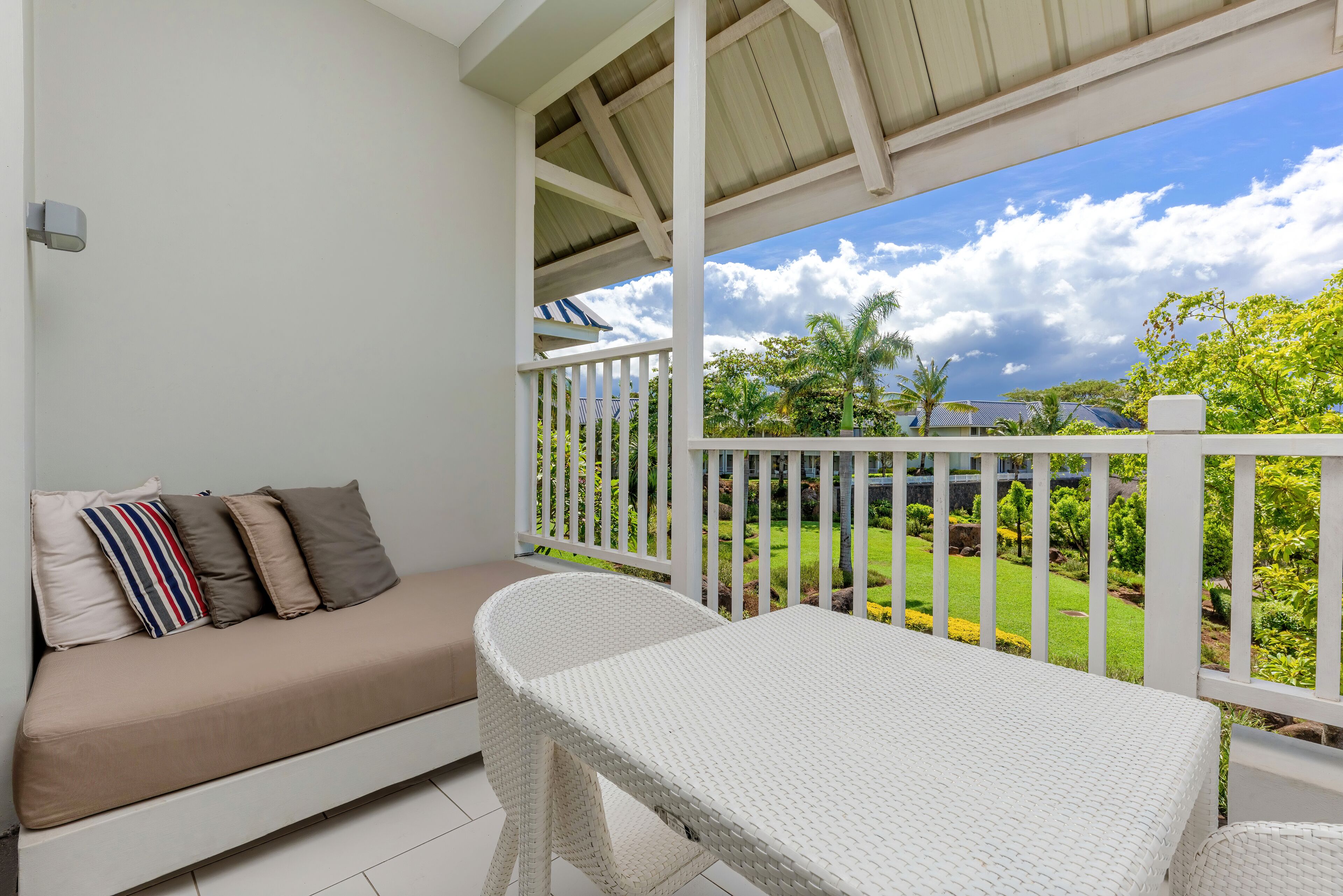 A balcony with a white wicker sofa and a table, overlooking a well-maintained garden and a clear sky.