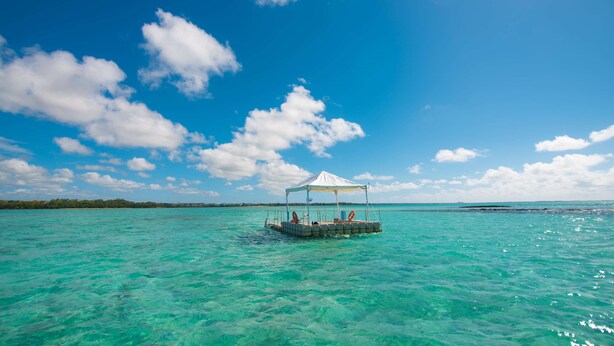 Una plataforma flotante con toldo, rodeada de agua turquesa cristalina y una costa lejana.