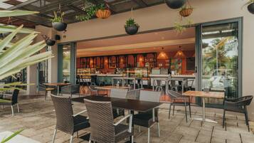Un café al aire libre con mobiliario de mimbre, plantas colgantes y vistas al área del bar interior.