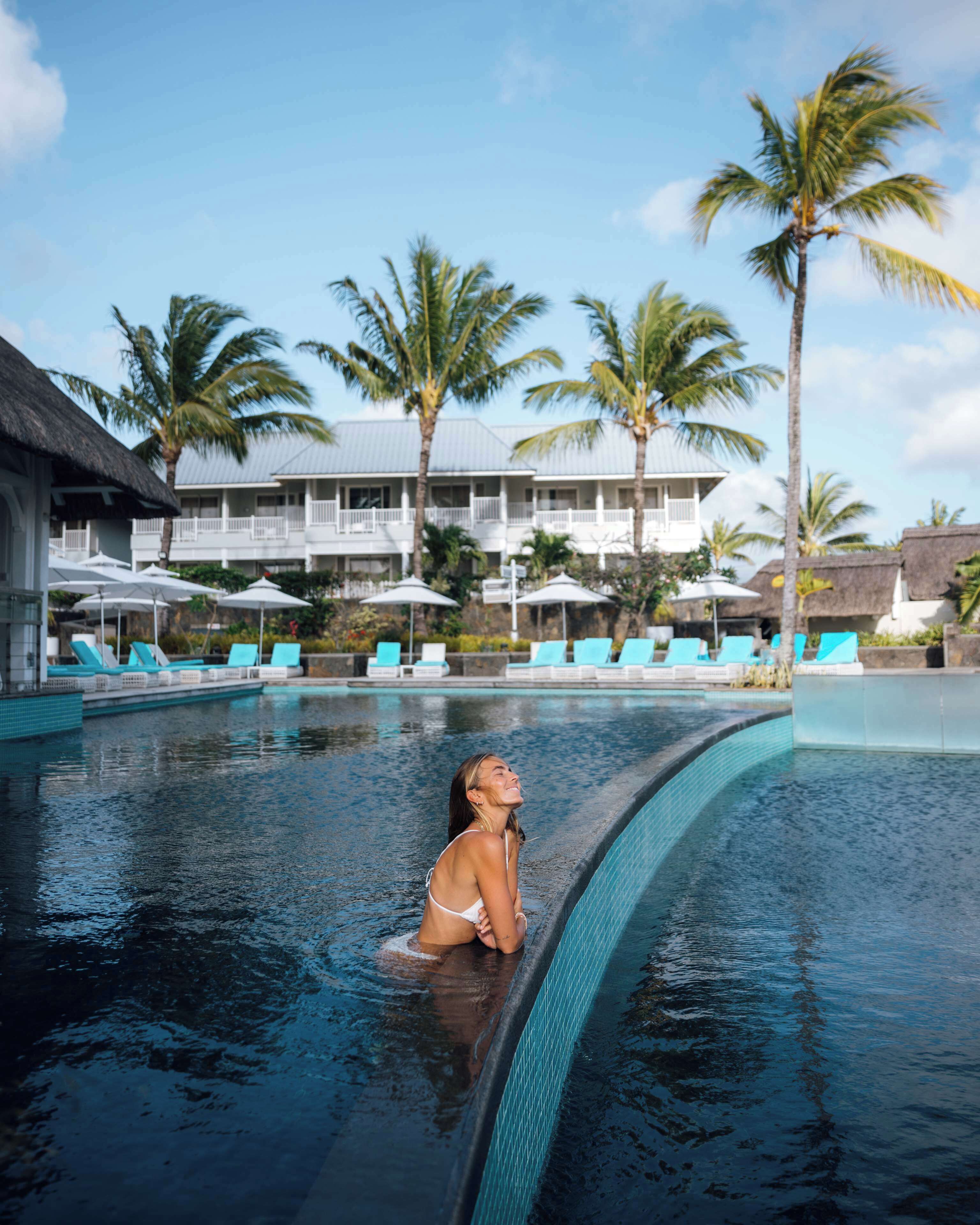Un espace piscine avec des chaises longues et des parasols, des palmiers et, au fond, un bâtiment doté de balcons.