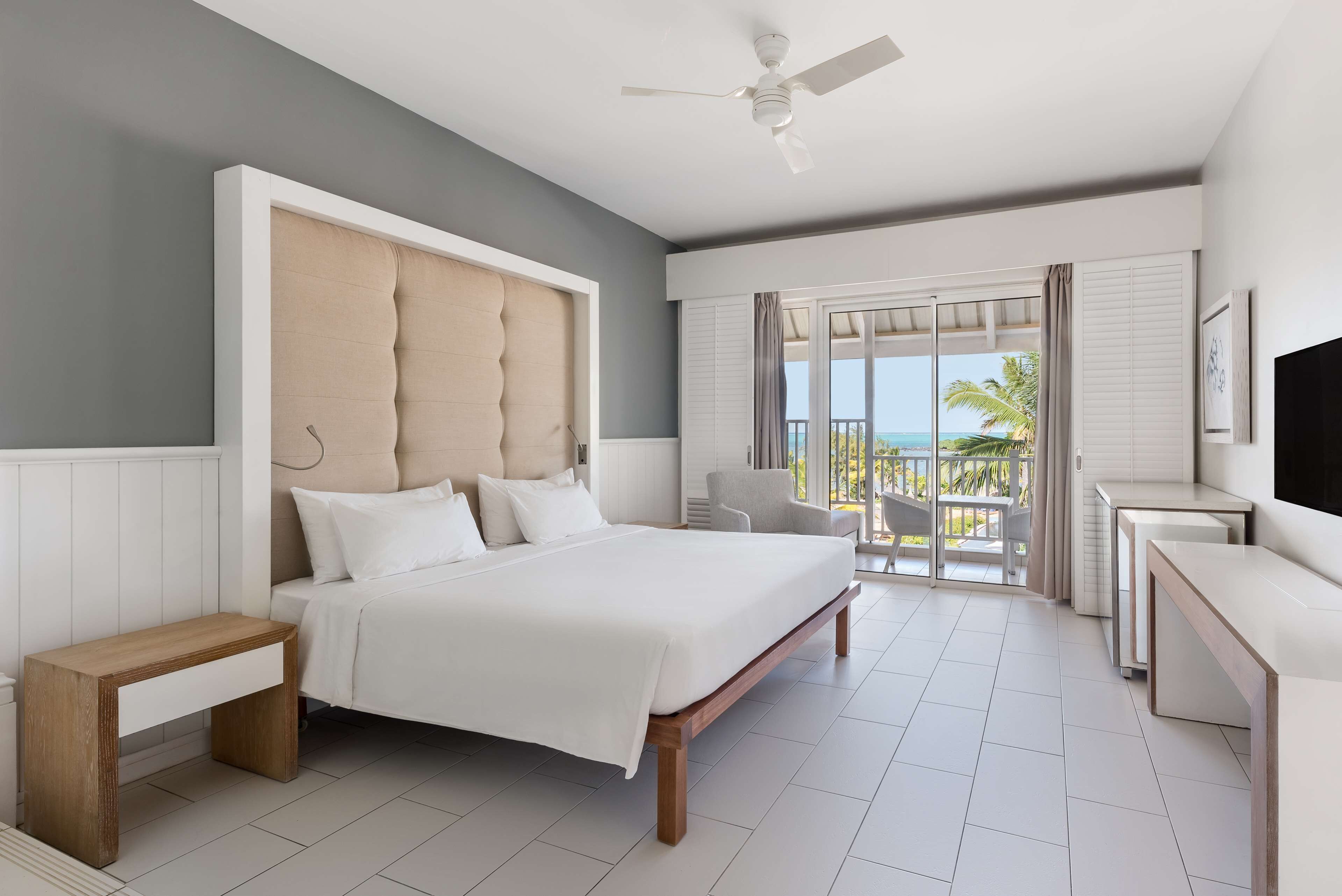 A modern hotel room with a large bed, a ceiling fan, a balcony with a view of the ocean, and a desk with a chair.