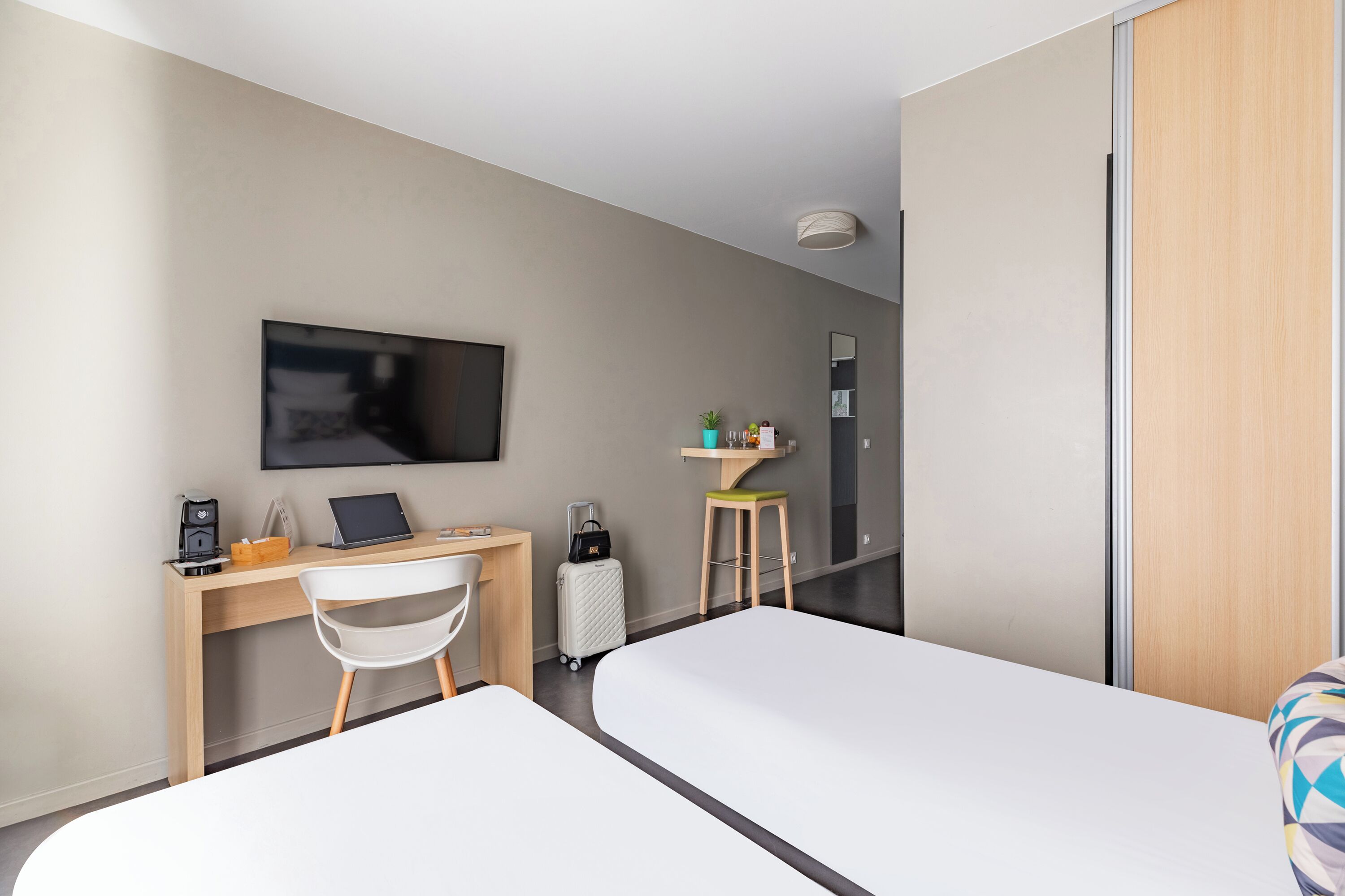 Ein Hotelzimmer mit zwei Betten, einem Schreibtisch mit einem Laptop, einem Sessel, einem Fernseher, einem kleinen Hocker und einem Koffer.