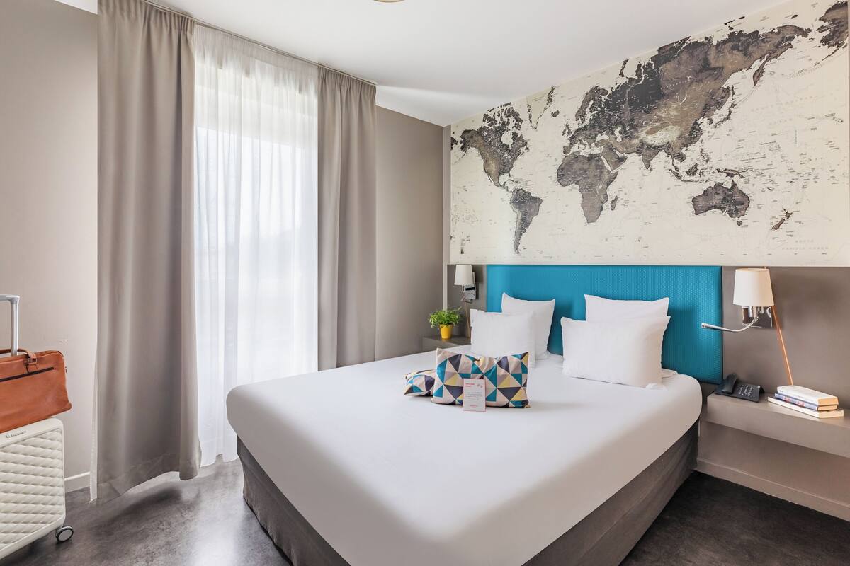 Een hotelkamer met een groot bed, een wereldkaart aan de muur, een blauw hoofdeinde, een nachtkastje met een lamp en een koffer op de vloer.