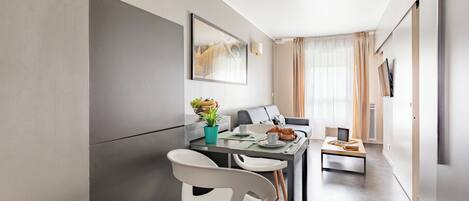 A modern, minimalist dining area with a table set for two, a sofa, and a TV in the background.