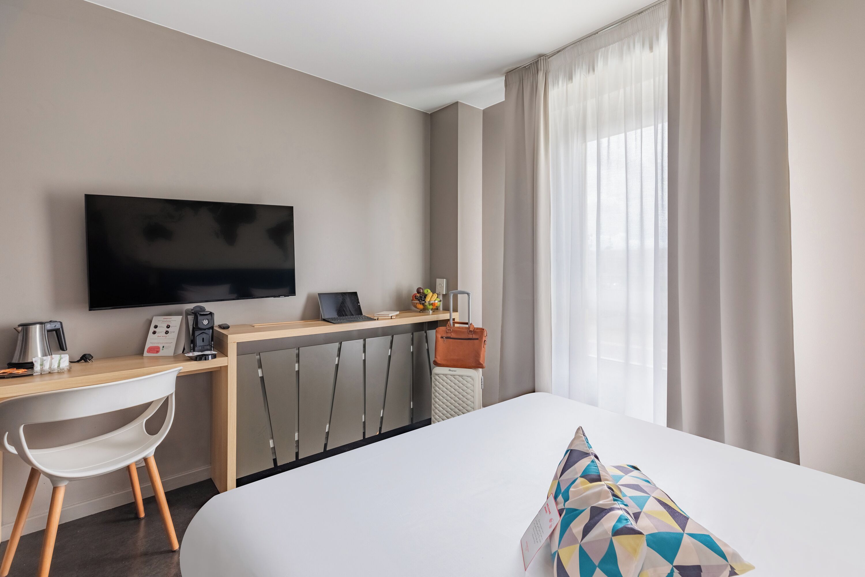 Habitación de hotel con una cama, un escritorio con televisor, una silla, una maleta y una ventana con cortinas.