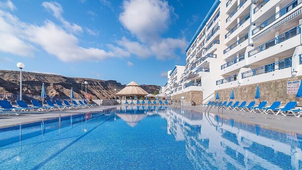 A swimming pool with lounge chairs and a building with balconies. - HV Apartamentos Vista Taurito (Mogan)