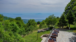 A patio area with stone flooring and wooden furniture, overlooking a scenic landscape with trees and a distant view of mountains and a body of water.