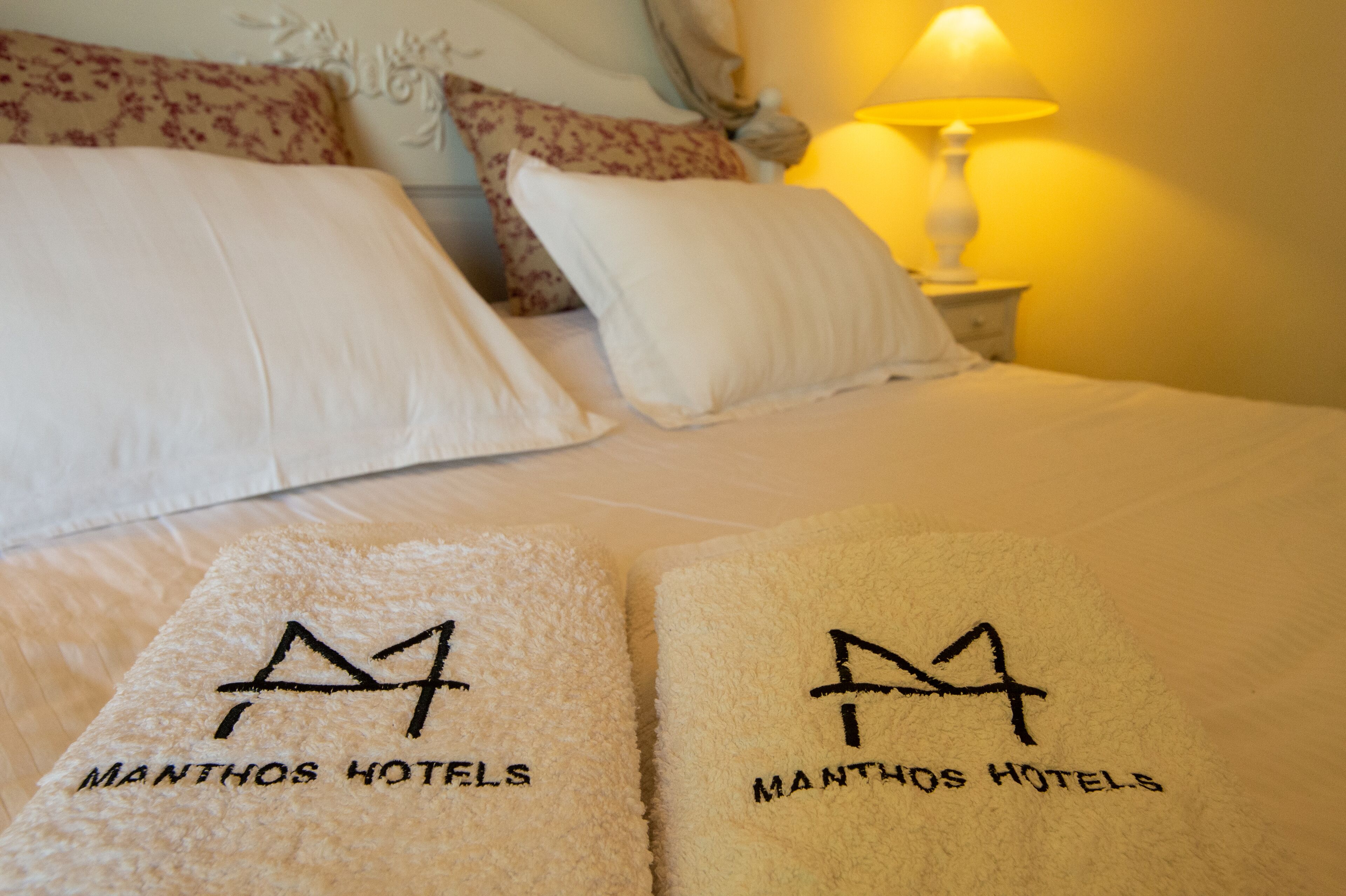 A bed with white linens and two towels with 'Manthos Hotels' branding.