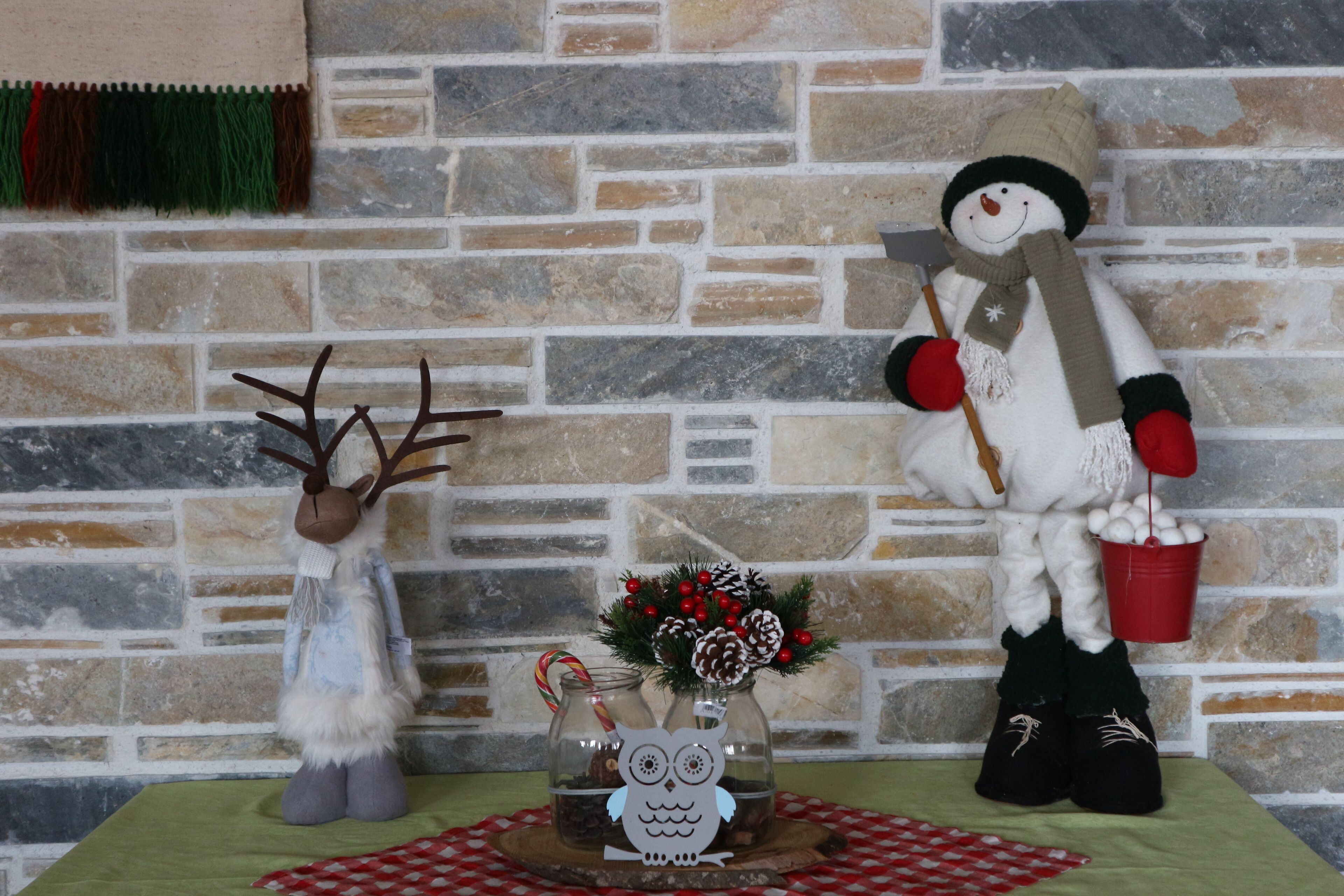 A rustic stone wall with a decorative glass vase containing pinecones and red berries, a plush reindeer, and a snowman figure holding a bucket of eggs.