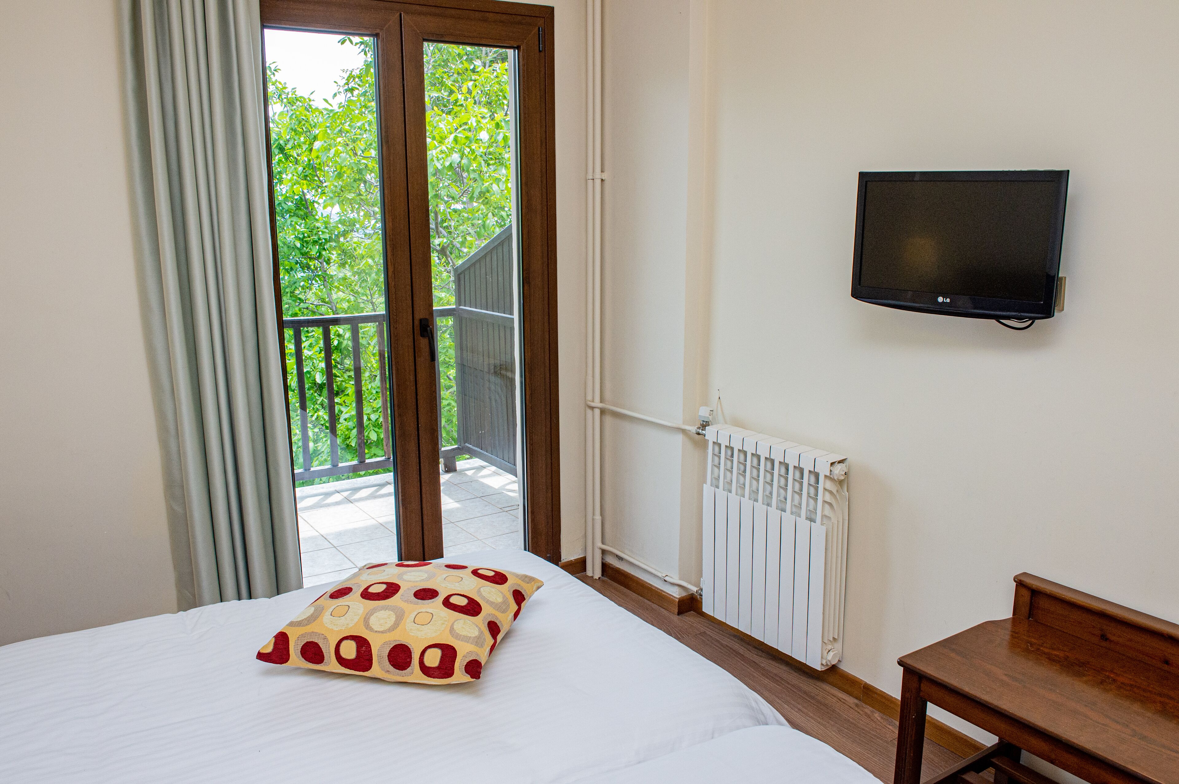 Een hotelkamer met een bed, een aan de muur bevestigde televisie, een raam met gordijnen en uitzicht op een balkon met een reling.
