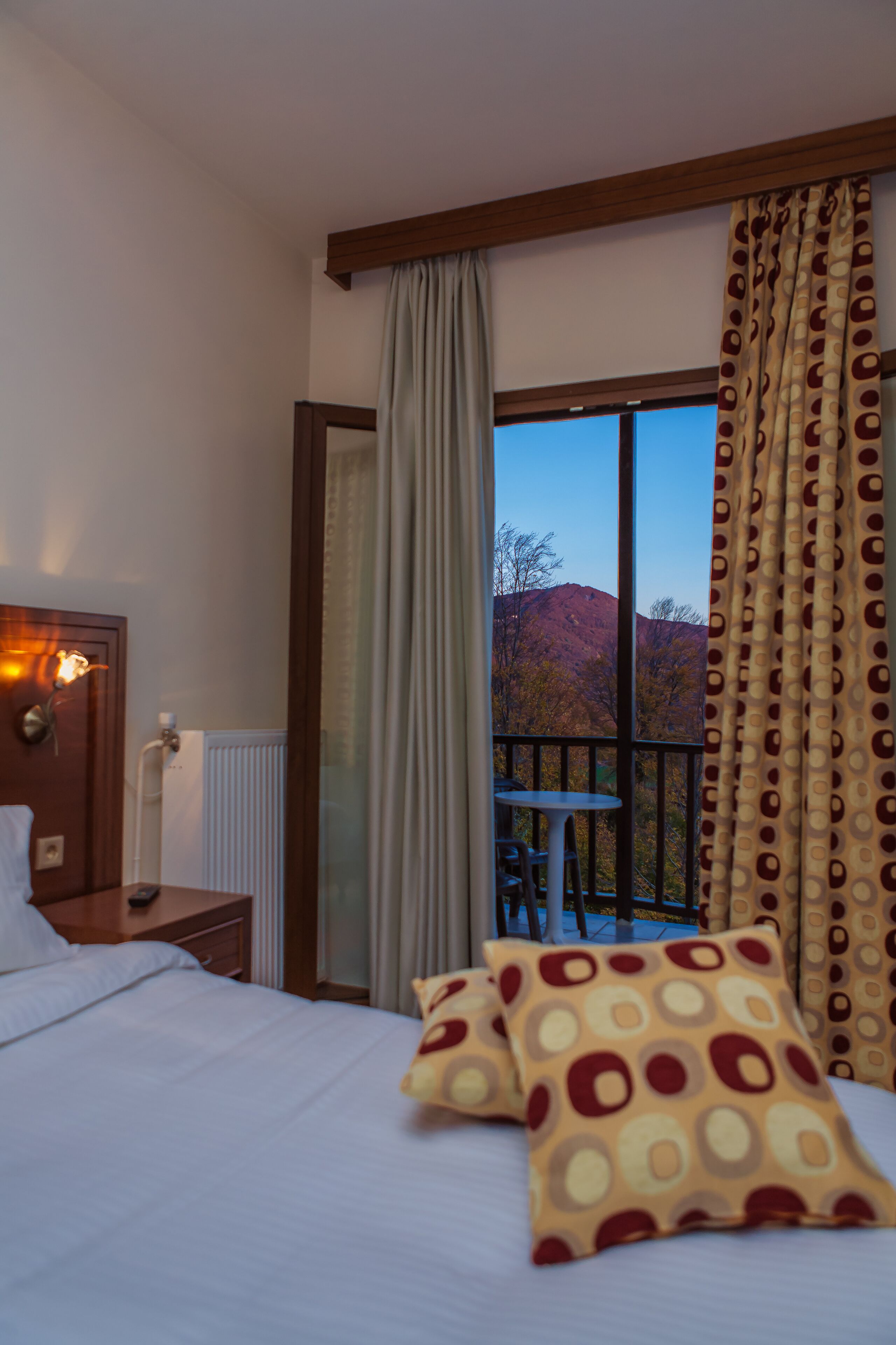 A hotel room with a bed, a nightstand, a lamp, a window with curtains, and a view of a mountain.