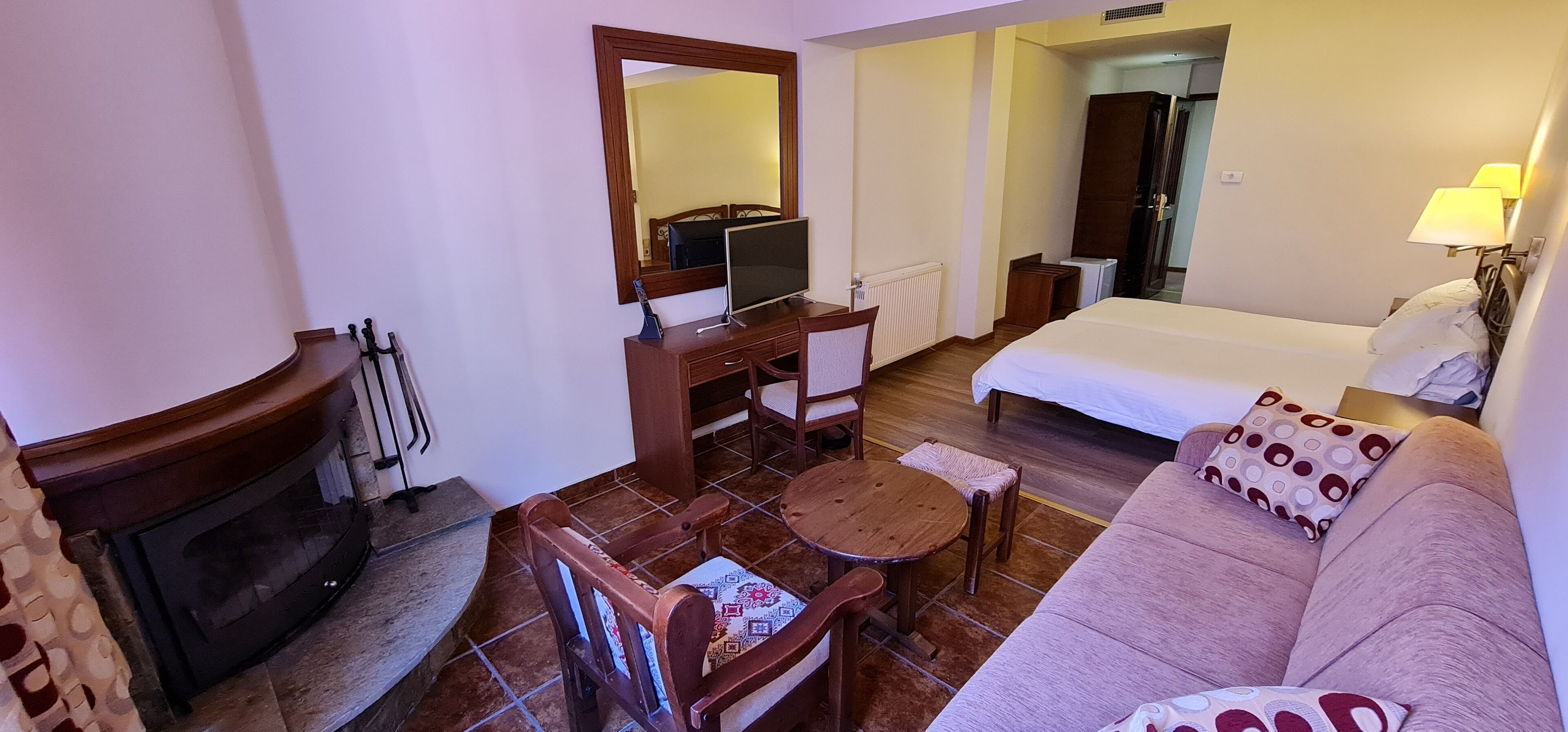 A hotel room with a bed, a desk with a computer, a chair, a small table, a fireplace, and a sofa.