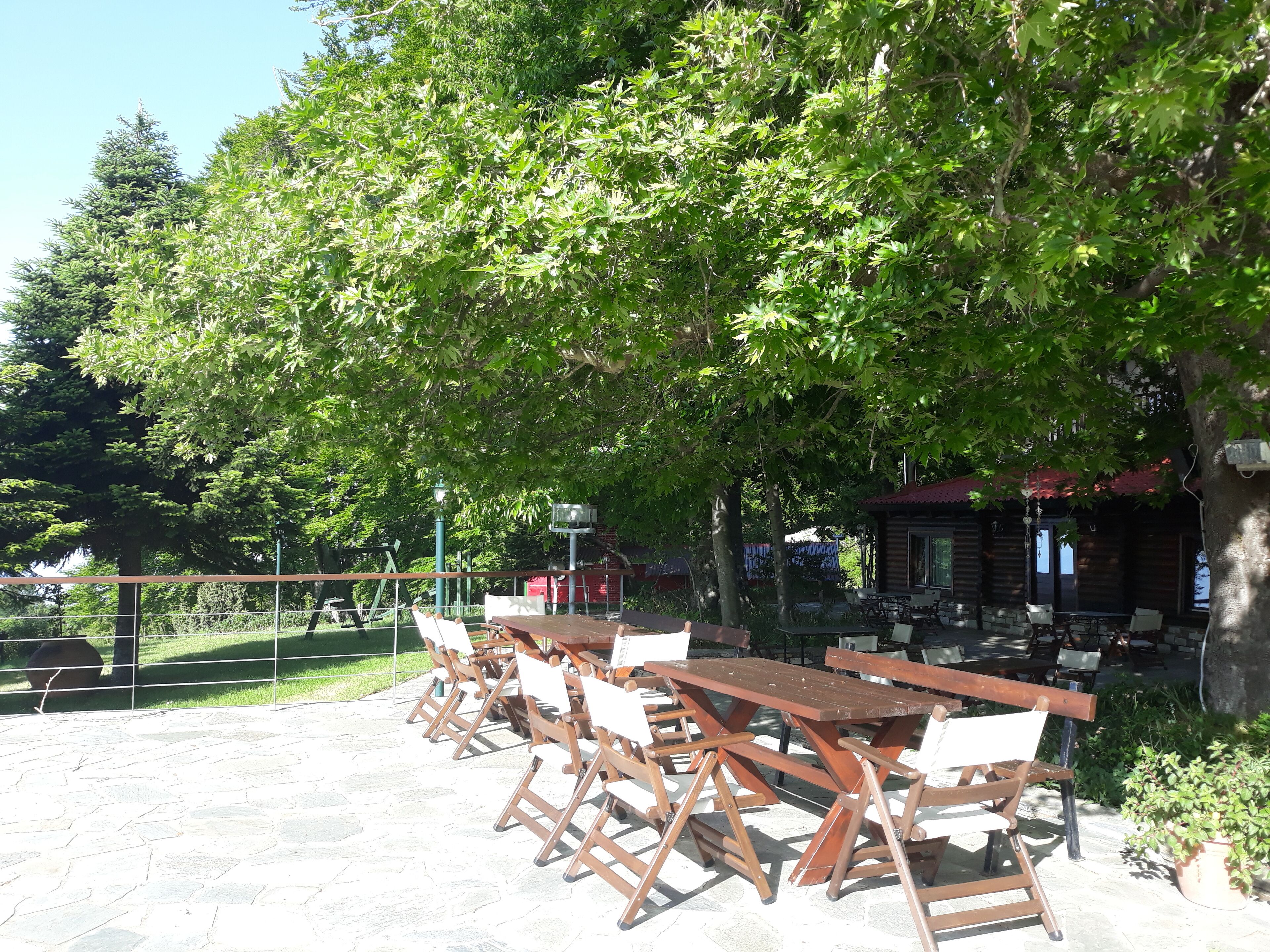 An outdoor seating area with tables and chairs, surrounded by trees and a building with a red roof.