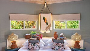 A room with a bed, two side tables, a hanging light fixture, and windows with a view of greenery. - East Winds Saint Lucia (Gros Islet)