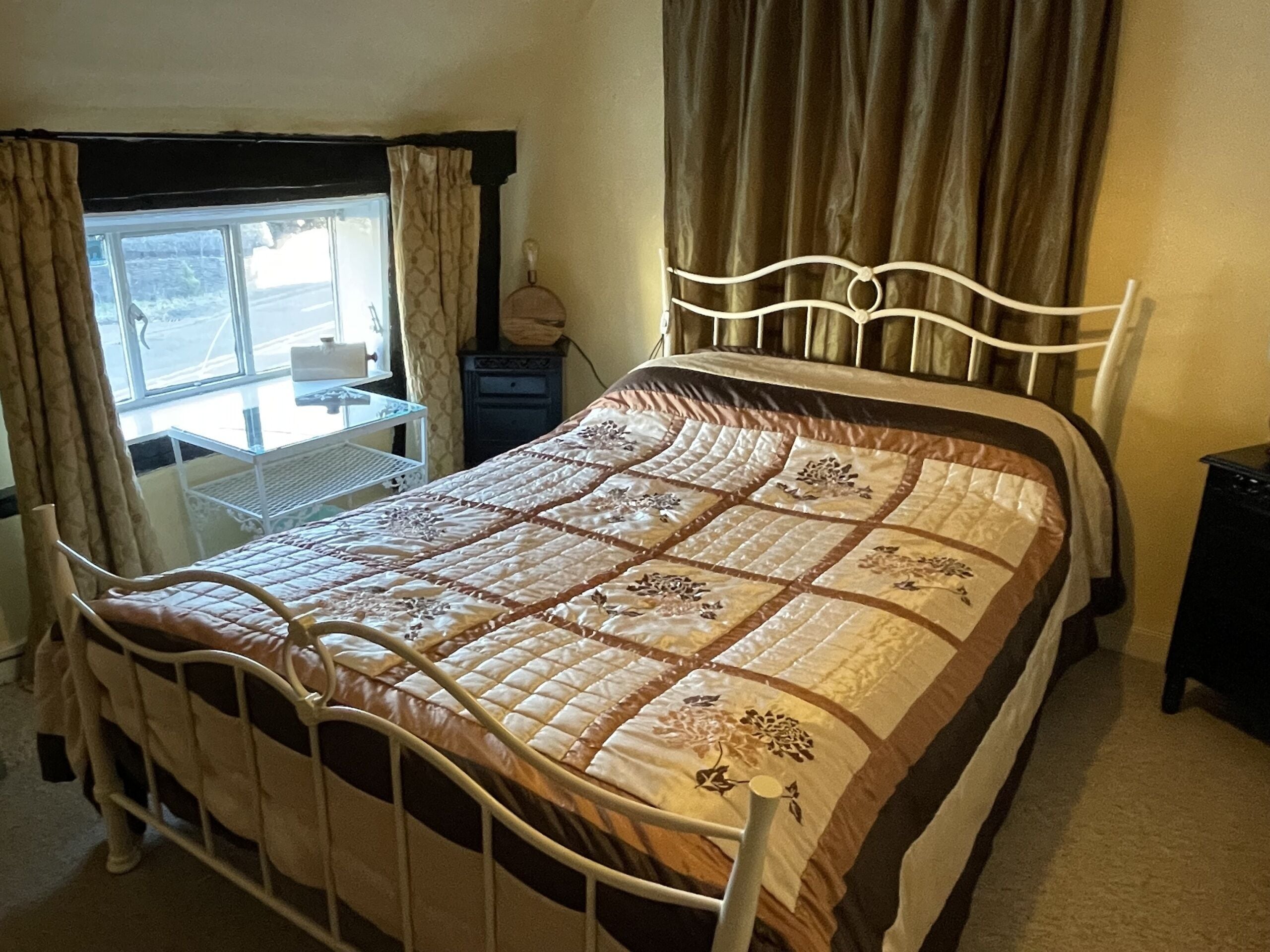 Een enkel bed met metalen frame, een quilt met bloemenmotieven, een nachtkastje en een raam met gordijnen.
