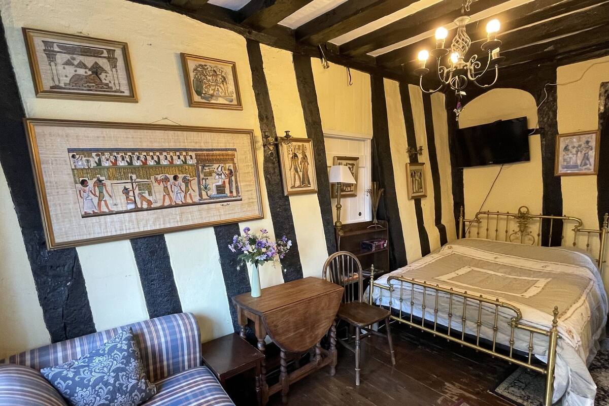 A traditional room with a bed, a wooden chair, a small table, and a desk. There are framed pictures on the walls.