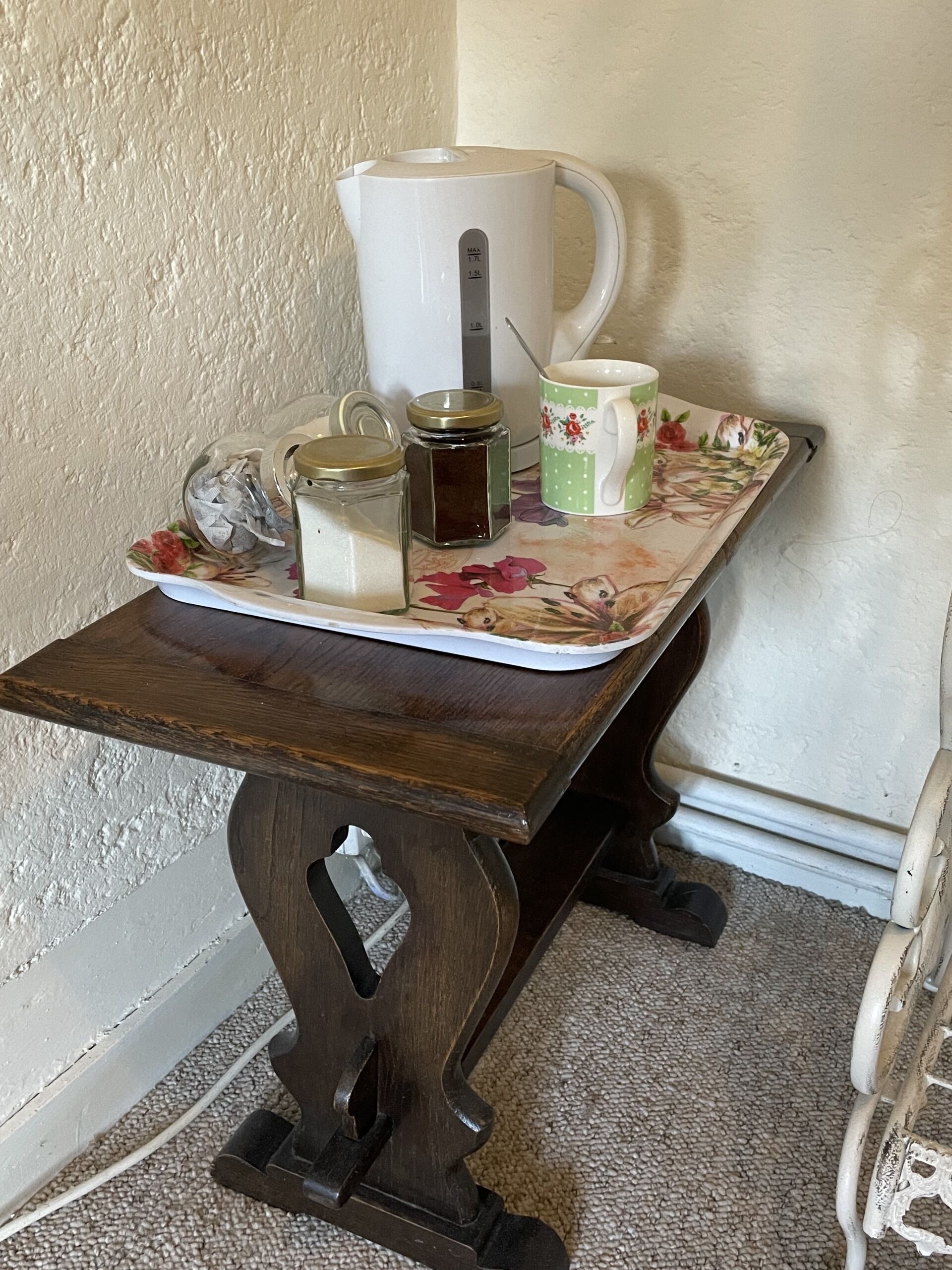 A small wooden table with a floral tablecloth, a white coffee pot, a green mug, two glass jars, and a metal container.