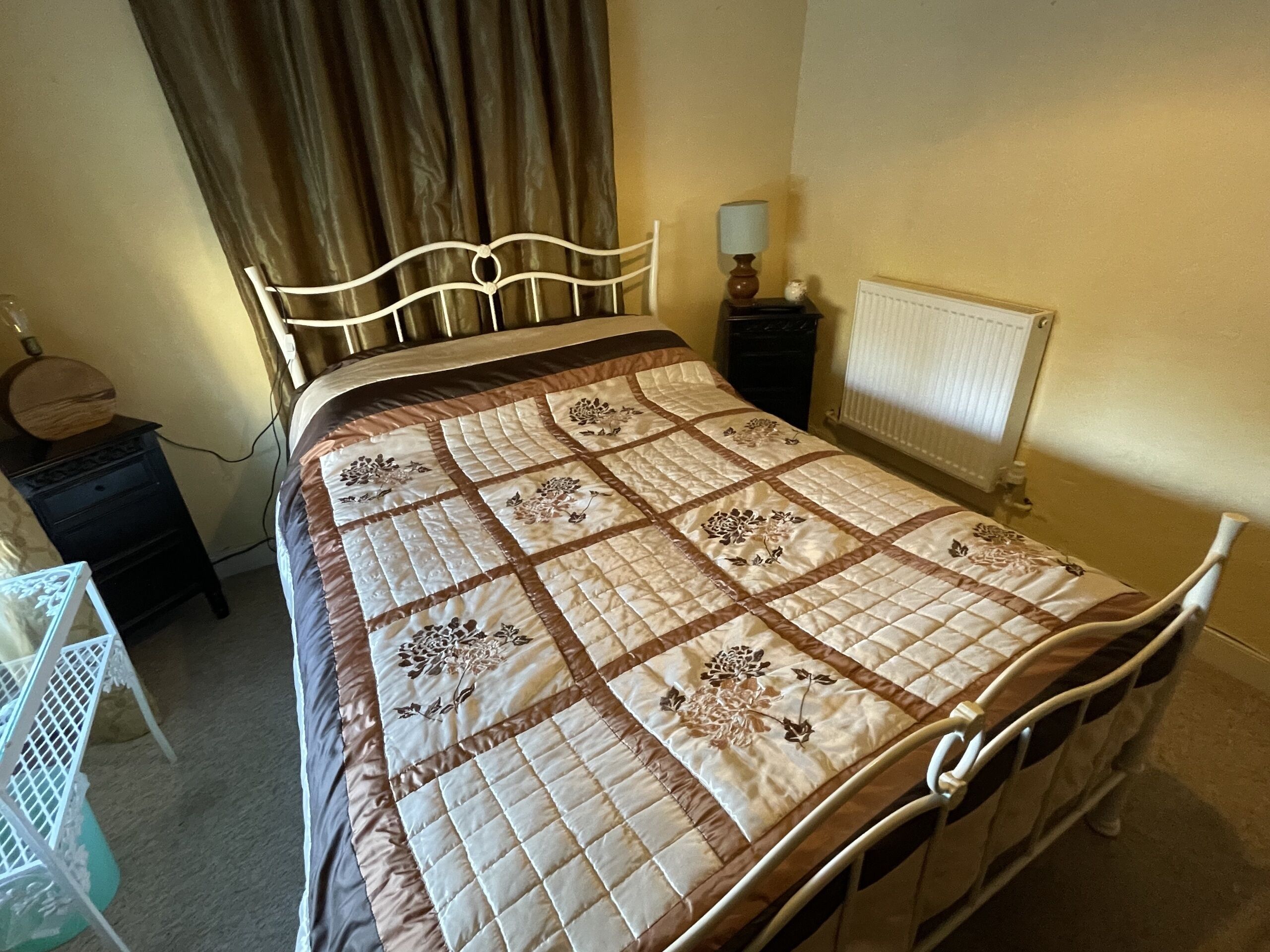 En pænt redt seng med et quiltet betræk, et sengebord med en lampe og en radiator på væggen.