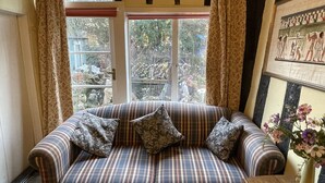 A striped sofa with patterned cushions, a wooden table with a vase of flowers, and a view of a garden through a window with curtains. - The Bridge Street Historic Guest House (Sudbury)