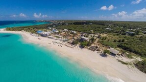 An aerial view of a coastal resort with a sandy beach, clear turquoise waters, and a mix of buildings and greenery.