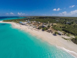 An aerial view of a coastal resort with a sandy beach, clear turquoise waters, and a mix of buildings and greenery.